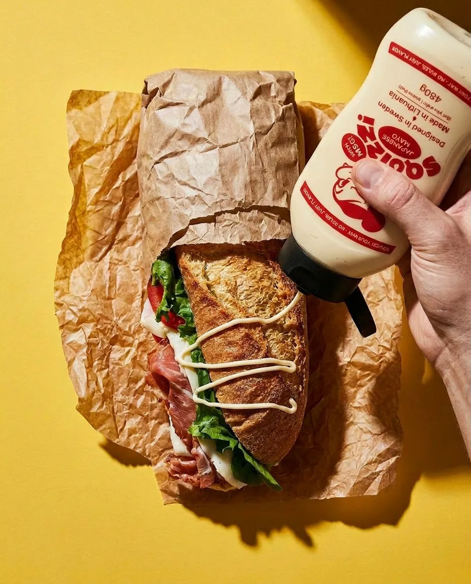 Hand drizzling Squizzi mayo onto a baguette over a yellow backdrop
